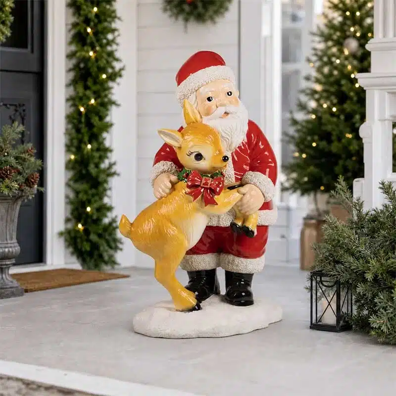 Santa-Standing-with-Reindeer-Christmas-Decoration Standing Santa holding a young reindeer with a red bow on a snow-effect base